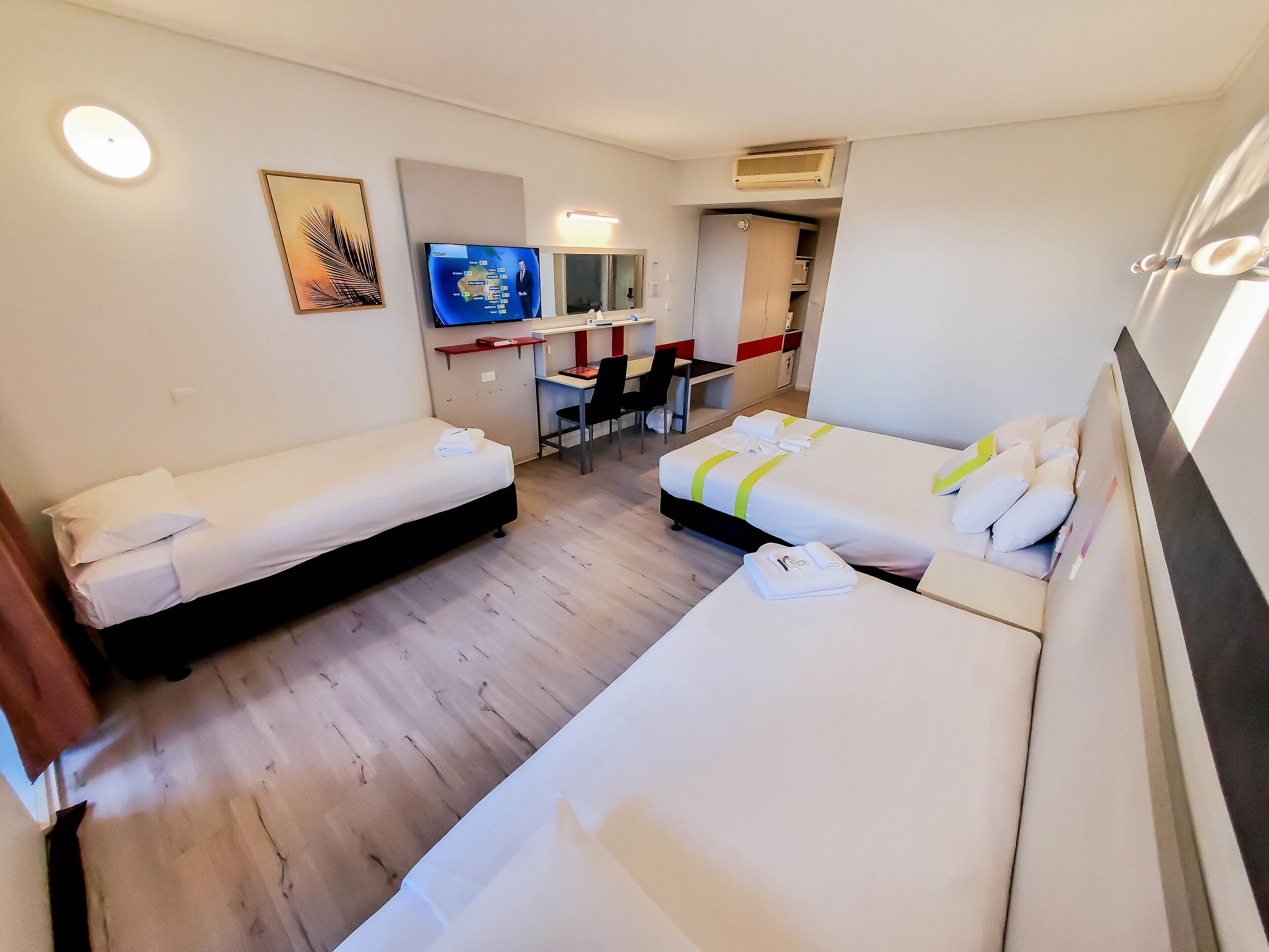 A hotel room with two single beds, a desk, a chair, a TV, and a small kitchenette.