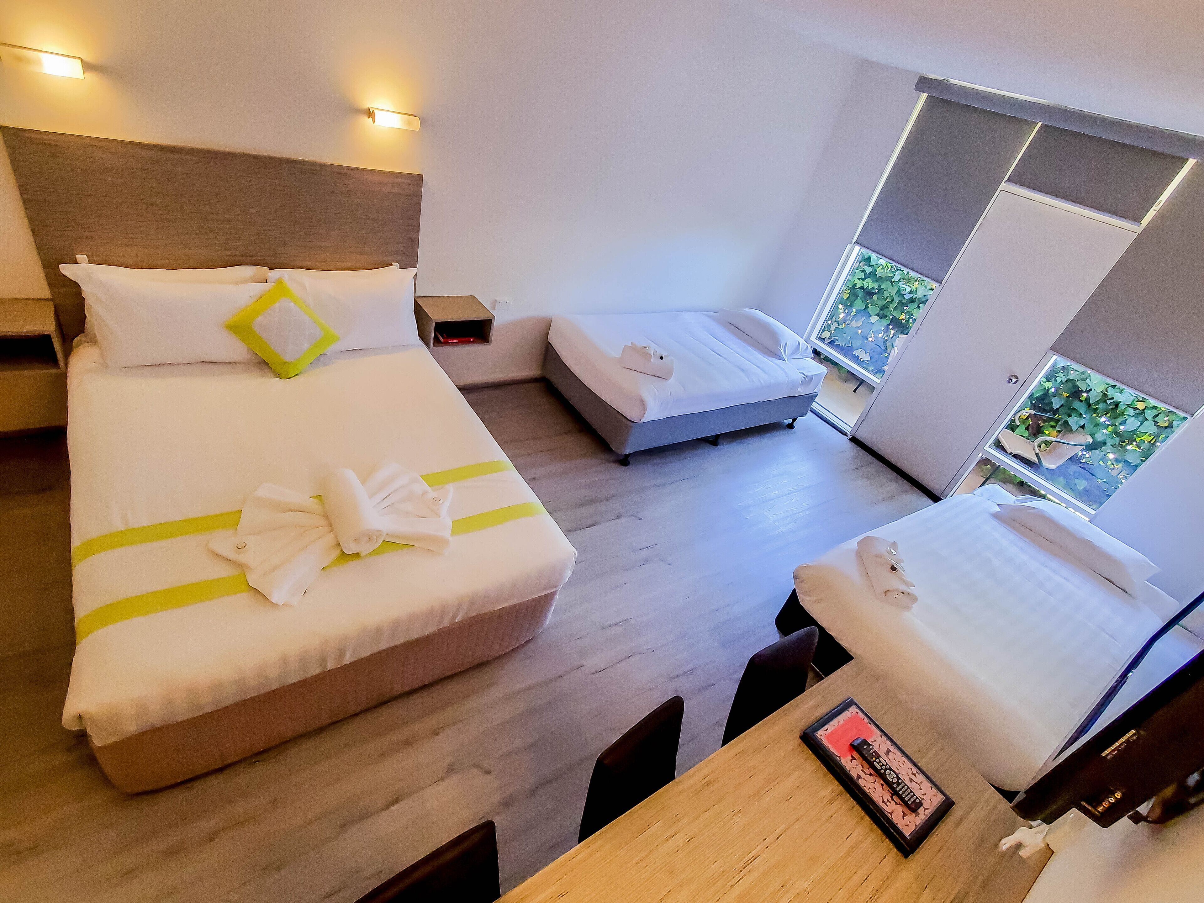 Hotelkamer met twee bedden, een houten vloer, een tafel met een afstandsbediening en een raam met uitzicht op groen.