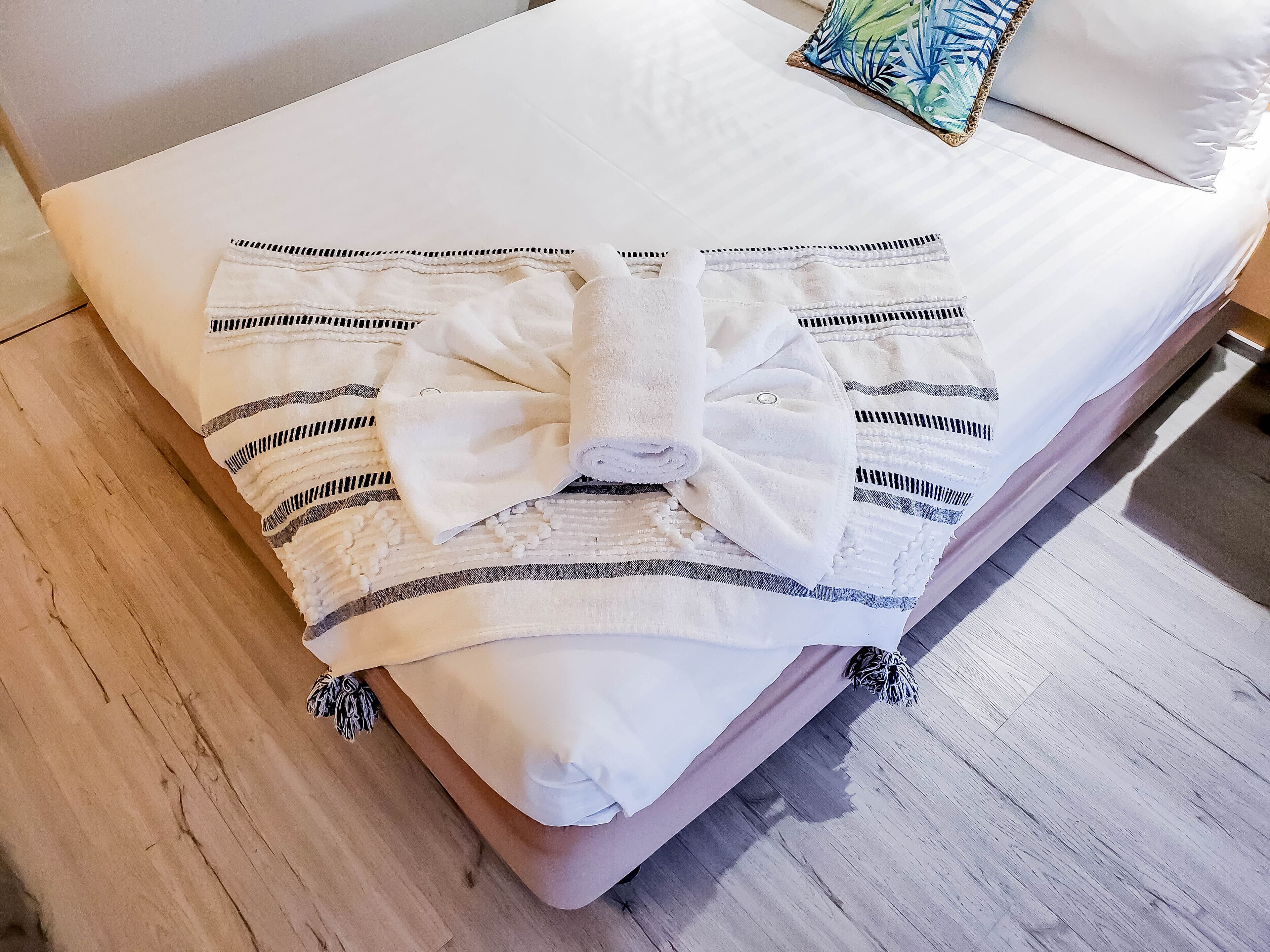 A neatly made bed with a white pillow and a decorative bow, a wooden floor, and a colorful pillow on the headboard.