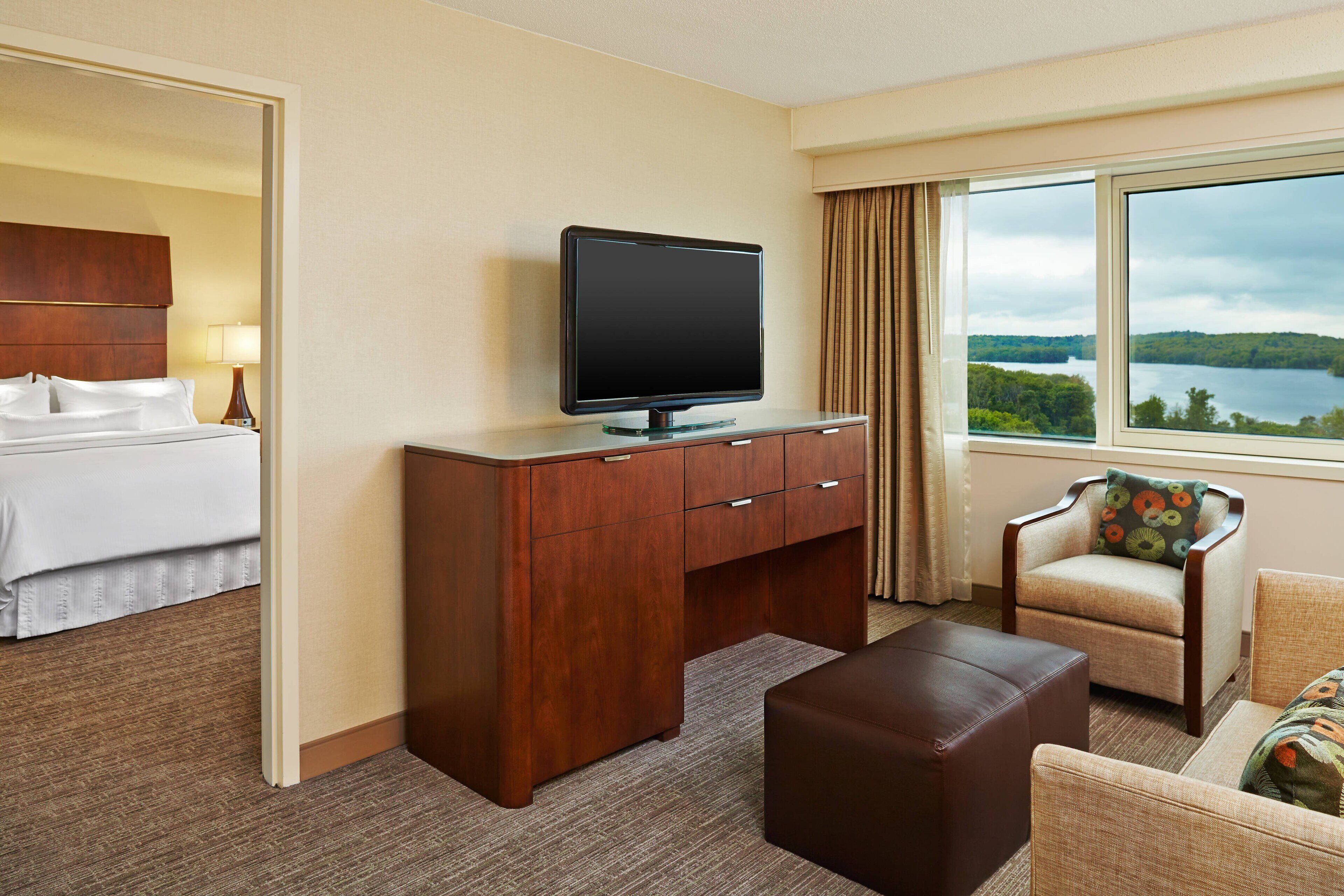 A hotel room with a bed, a wooden desk with a TV, a comfortable armchair, and a large window with a lake view.