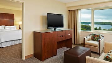 A hotel room with a bed, a wooden desk with a TV, a comfortable armchair, and a large window with a lake view.