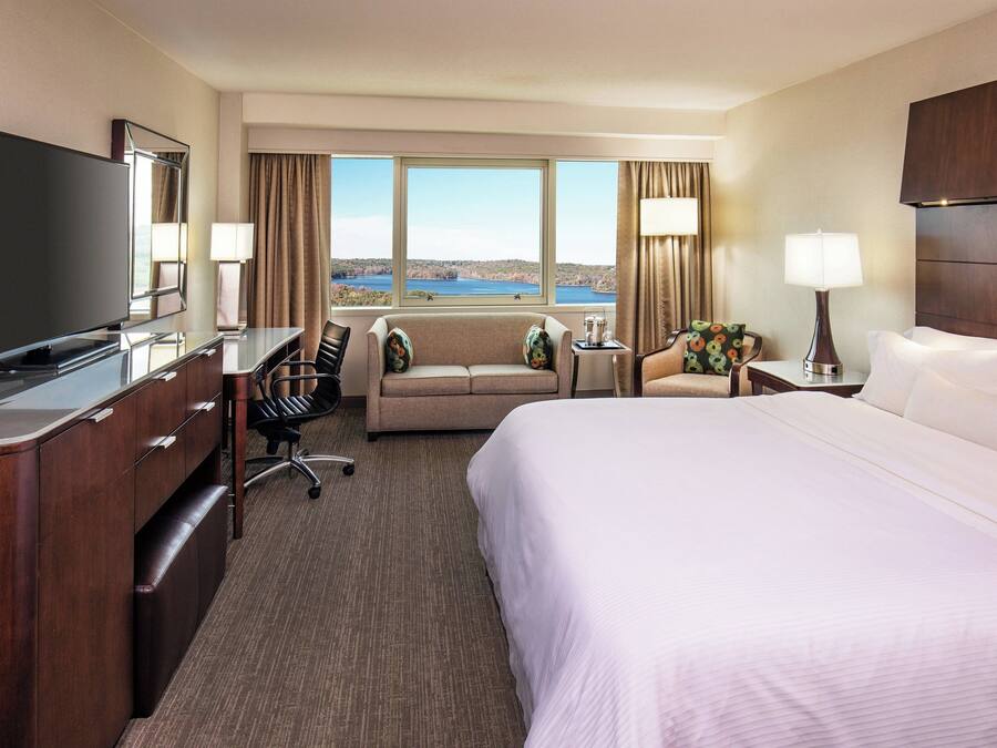 A hotel room with a large bed, a desk with a flat-screen TV, a sofa, a chair, and a view of a lake.