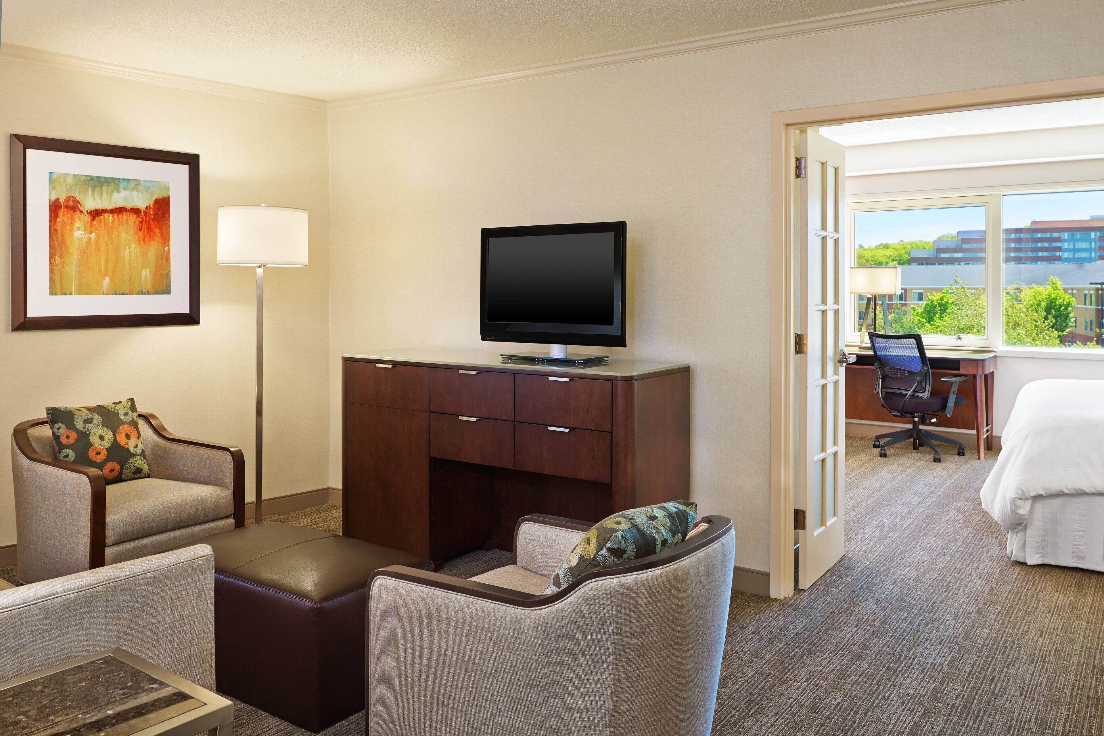 A hotel room with a television on a stand, a desk with a chair, a bed, and a window with a view of buildings and trees.