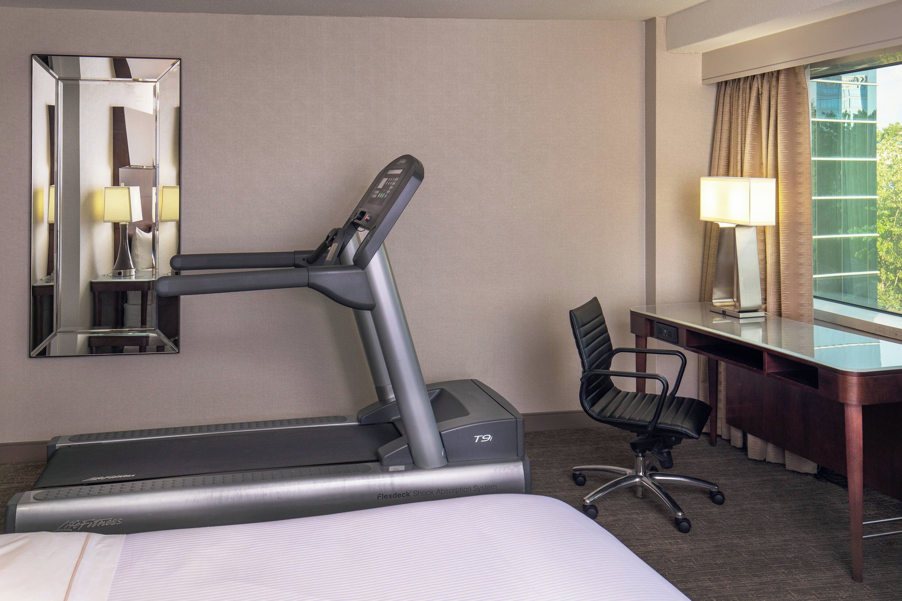 A hotel room with a treadmill, a desk with a chair, a large mirror, and a window with curtains.