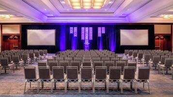 Un salón de conferencias con filas de sillas, un escenario con una pantalla grande y un telón de fondo azul oscuro.