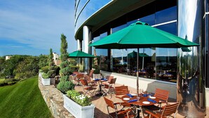 An outdoor patio with tables and chairs, green umbrellas, and a stone wall. - The Westin Waltham-Boston (Waltham)