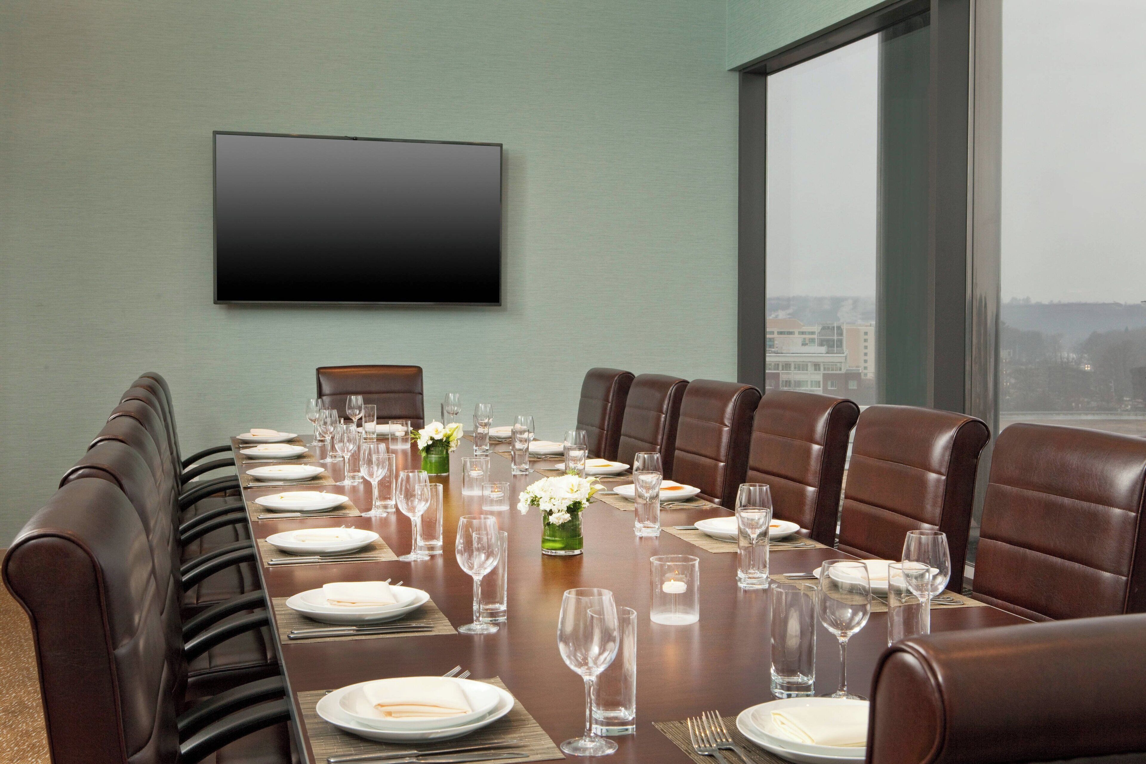 Un salón de conferencias con una larga mesa dispuesta para una reunión, sillas de cuero, un televisor de pantalla plana en la pared y vistas a la ciudad a través de grandes ventanales.