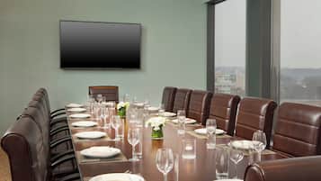 Un salón de conferencias con una larga mesa dispuesta para una reunión, sillas de cuero, un televisor de pantalla plana en la pared y vistas a la ciudad a través de grandes ventanales.