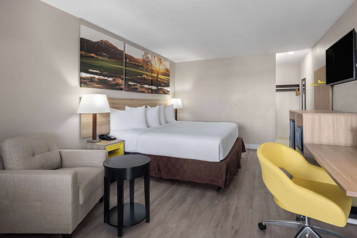 A hotel room with a bed, a desk, a chair, a small table, and a painting on the wall.