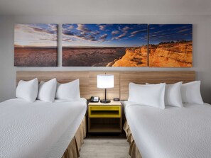 Quarto de hotel com duas camas, uma mesa de cabeceira com abajur e um quadro na parede retratando uma paisagem desértica.