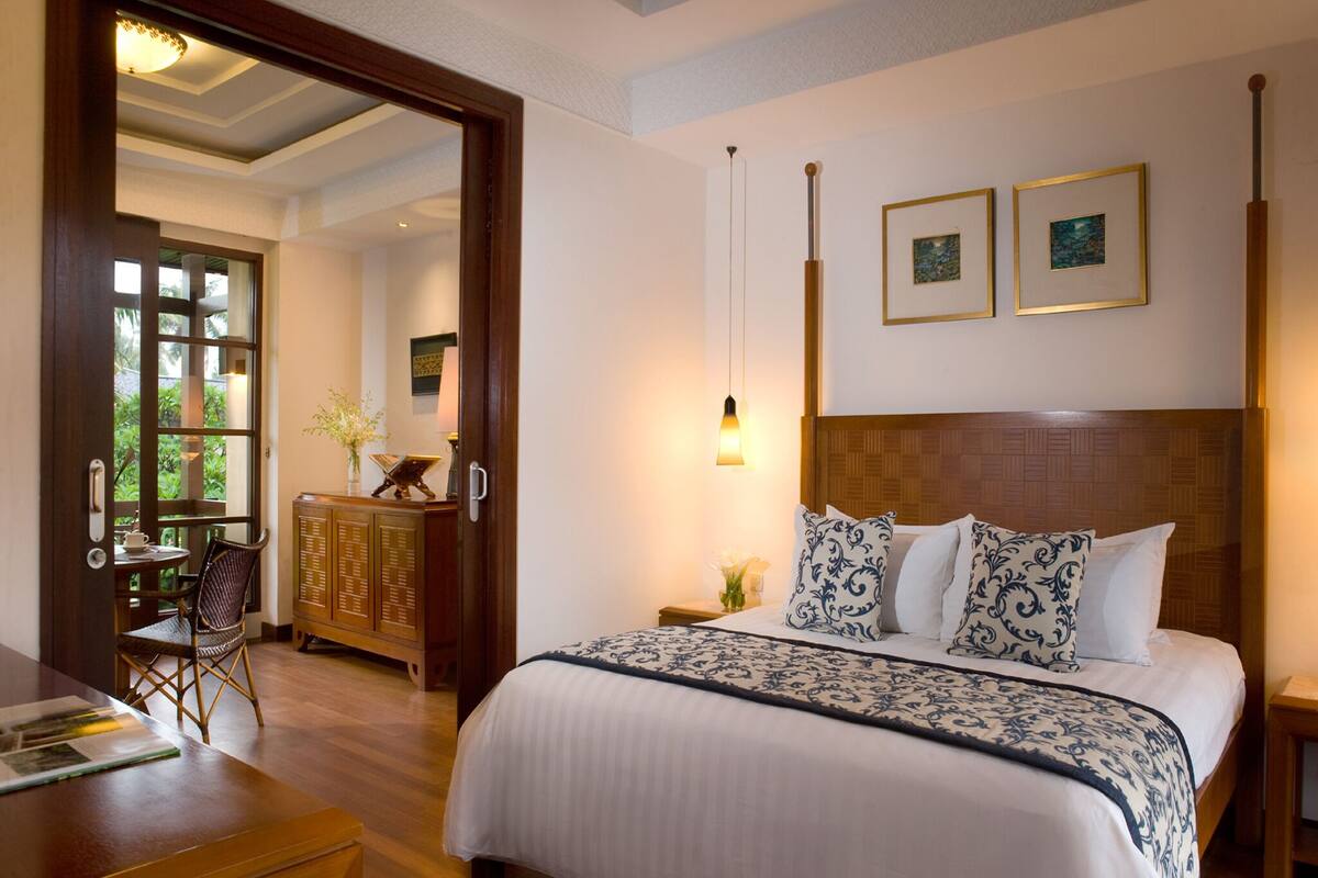 A hotel room with a large bed, a wooden headboard, a desk, and a chair. There are two framed pictures on the wall and a window with a view of greenery.
