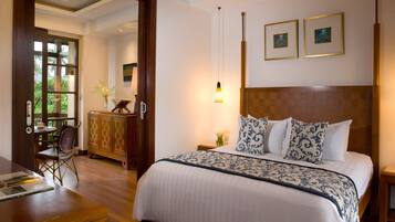 A hotel room with a large bed, a wooden headboard, a desk, and a chair. There are two framed pictures on the wall and a window with a view of greenery.