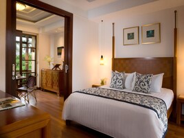 A hotel room with a large bed, a wooden headboard, a desk, and a chair. There are two framed pictures on the wall and a window with a view of greenery.