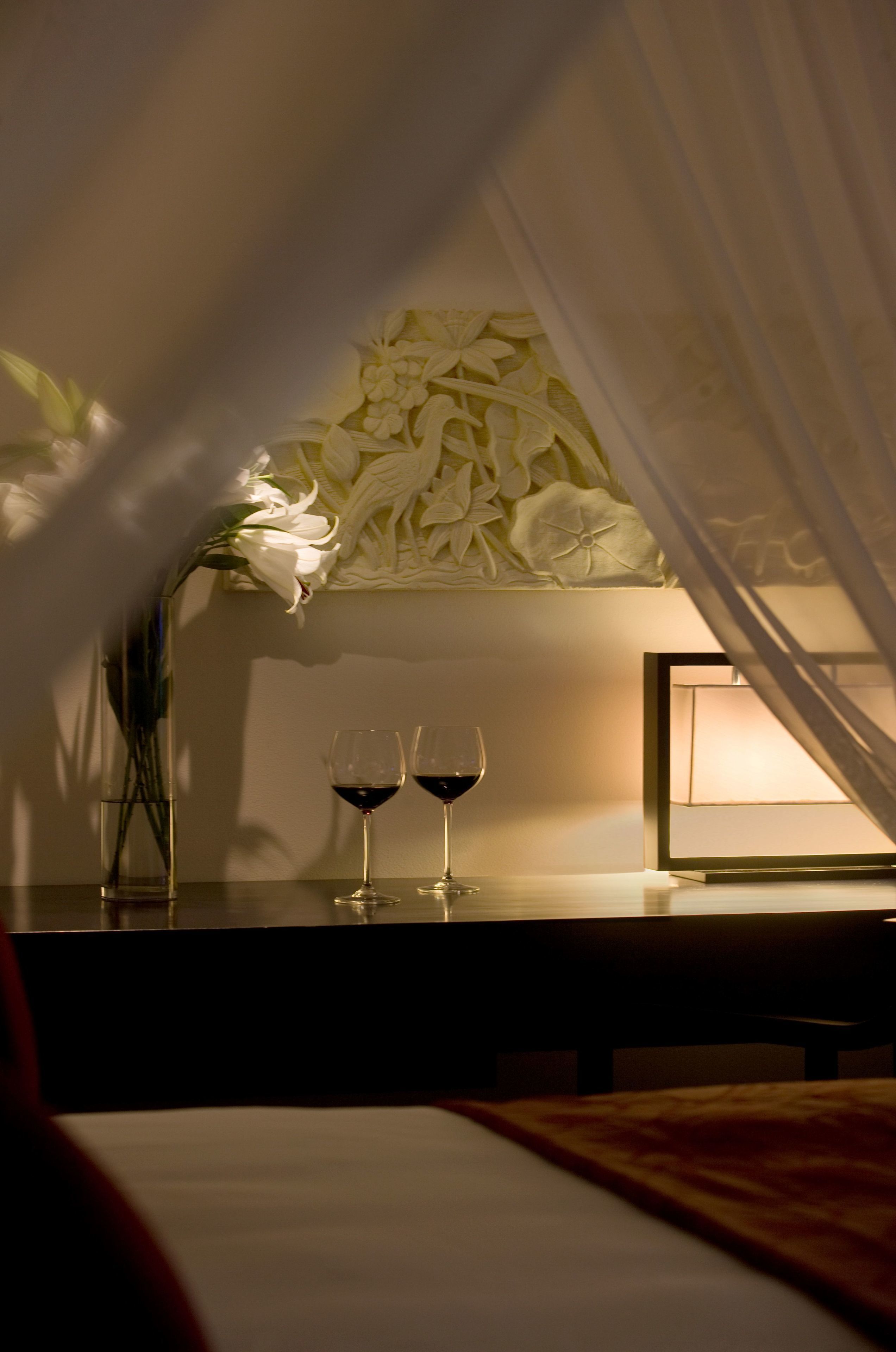 A dimly lit room with a bed, a bedside table with two wine glasses, a vase with flowers, and a wall with a floral pattern.