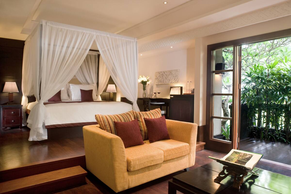 A bedroom with a four-poster bed, a sofa, a desk, and a view of greenery through a large window.
