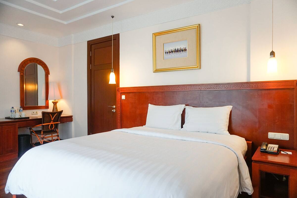 A hotel room with a large bed, a wooden headboard, a desk with a lamp, a mirror, and a framed picture on the wall.