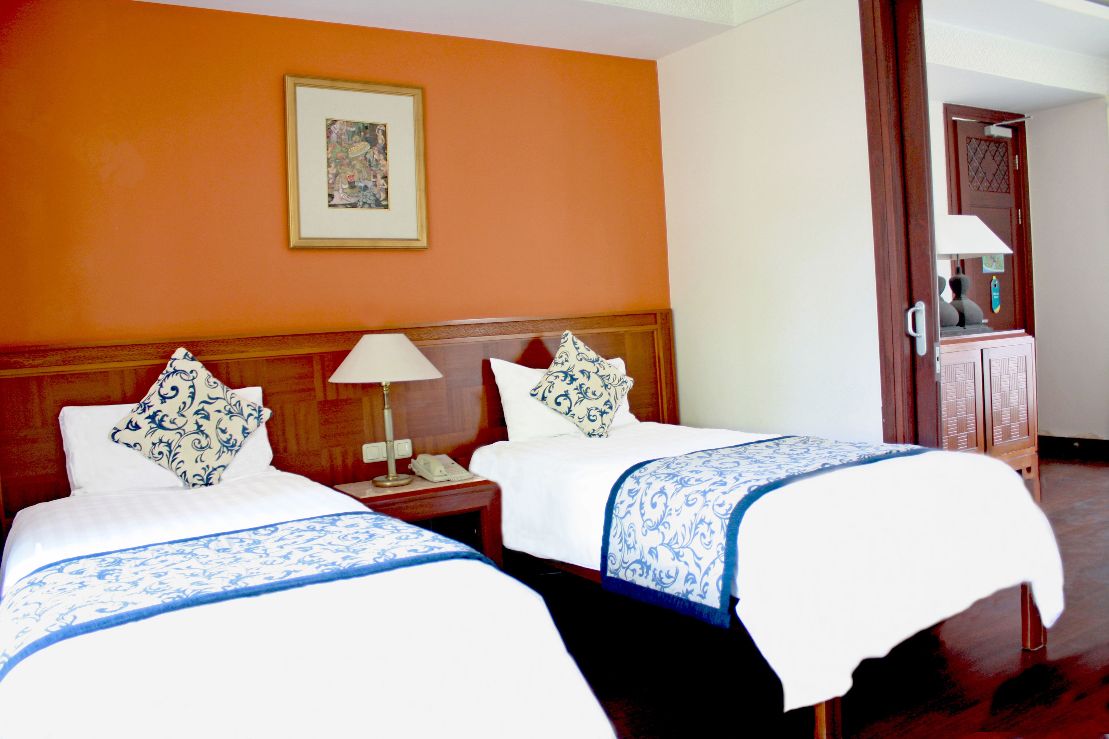A hotel room with two beds, a nightstand with a lamp, a framed picture on the wall, and a wooden door.