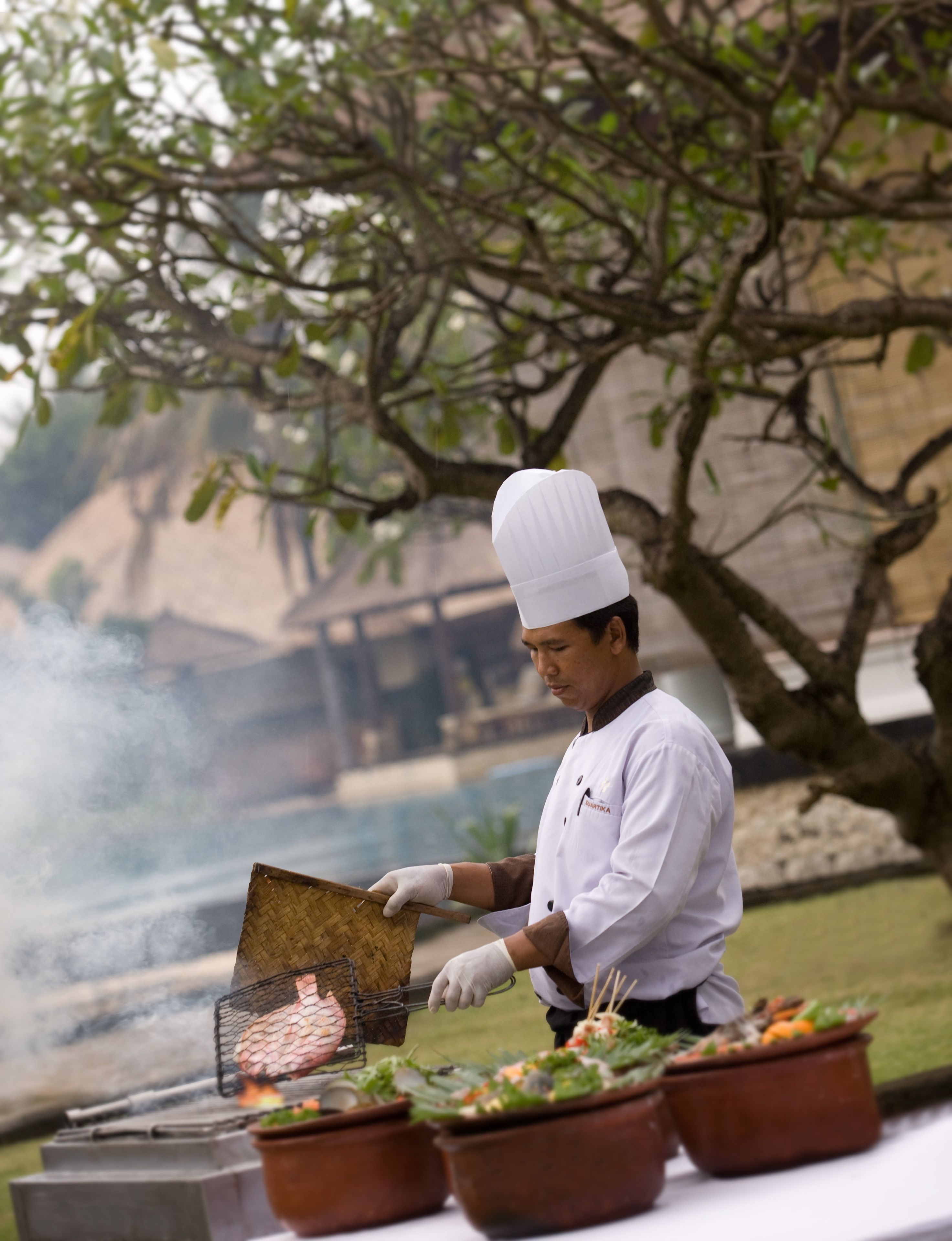 A chef grilling food outdoors with a view of a body of water and trees in the background.