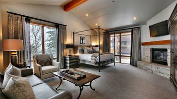 A spacious bedroom with a four-poster bed, a sitting area with a sofa and armchair, a fireplace, and a large window offering a view of the outdoors.