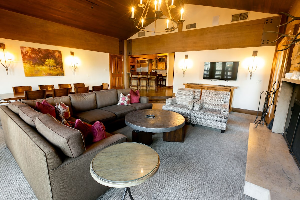 Ein geräumiges Wohnzimmer mit einer großen Eckcouch, einem runden Couchtisch und einem gemusterten Sessel. Ein Fernseher ist an der Wand montiert, und es gibt einen Kamin mit Steinverkleidung.