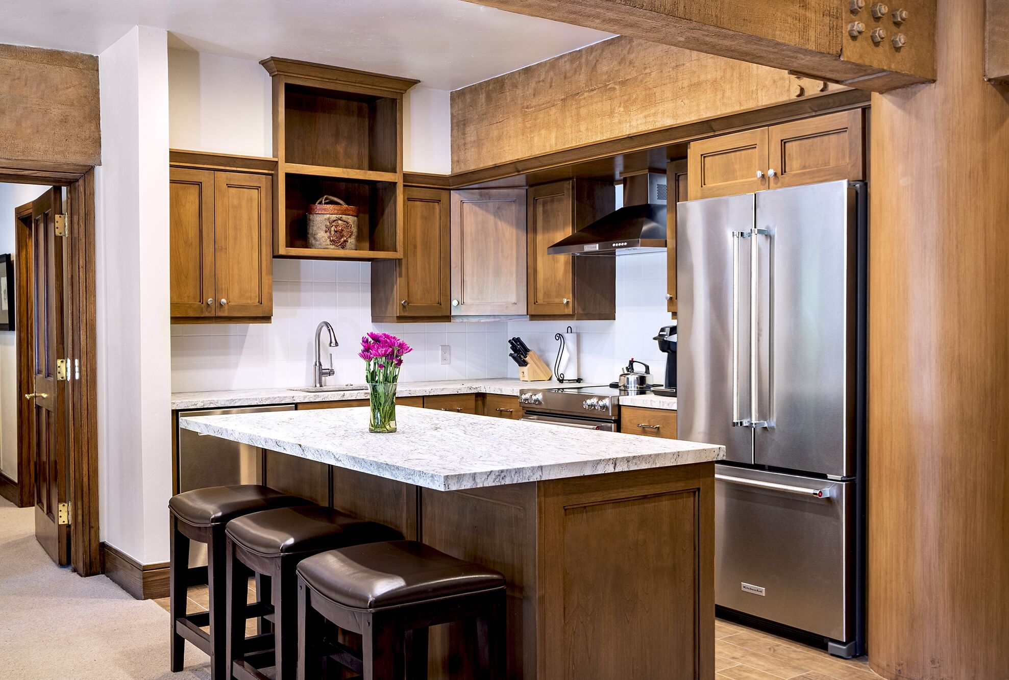 A kitchen with wooden cabinets, a stainless steel refrigerator, a marble countertop, and a vase with flowers.