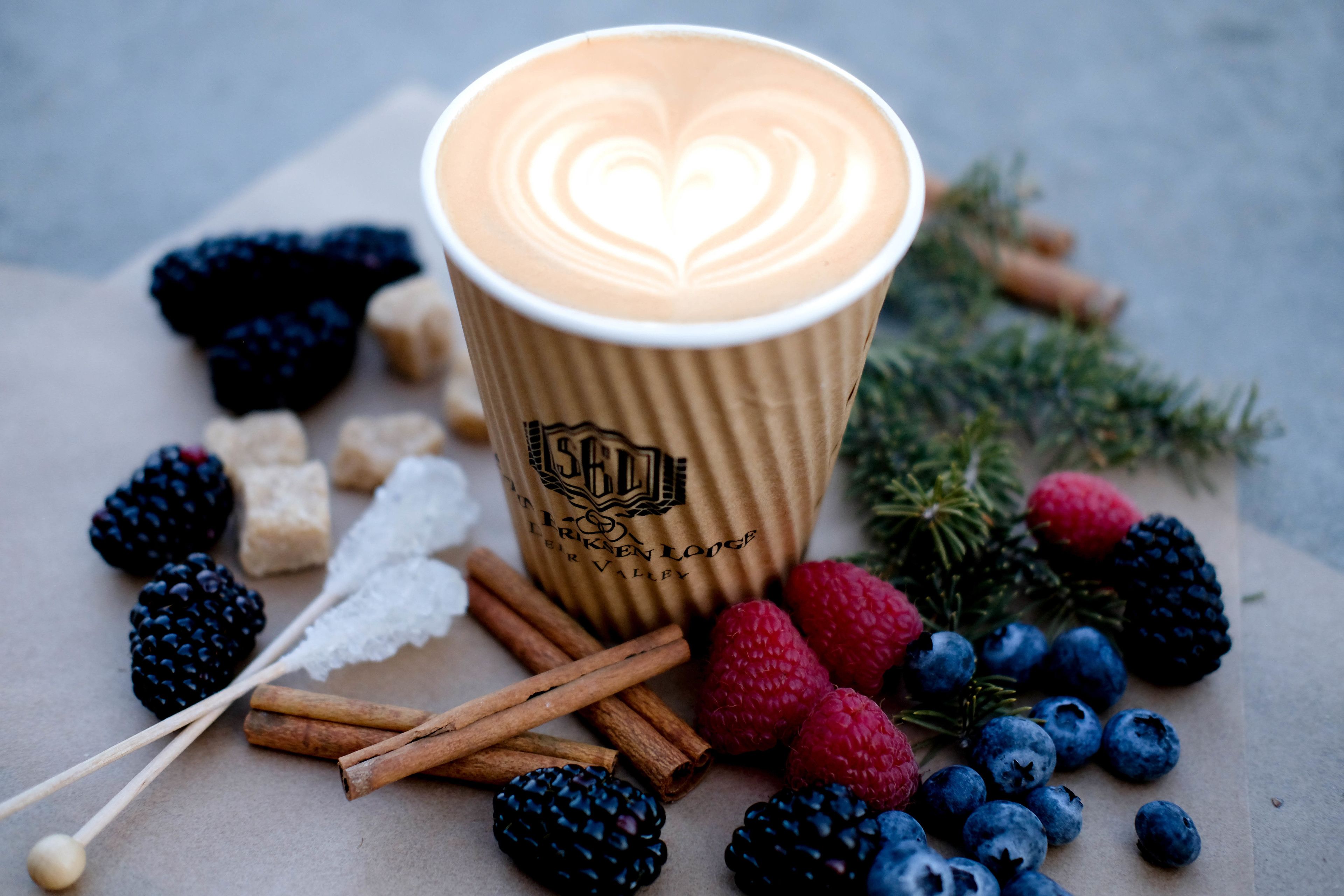 A cup of coffee with a heart design on the foam, surrounded by various berries and cinnamon sticks.