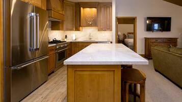A modern kitchen with wooden cabinets, a stainless steel refrigerator, a marble countertop, and a TV mounted on the wall.