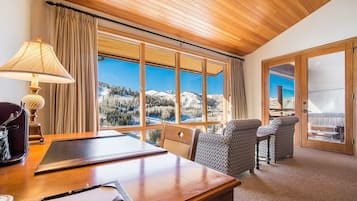 A room with a wooden desk, two patterned chairs, a lamp, and large windows offering a view of snow-covered mountains.