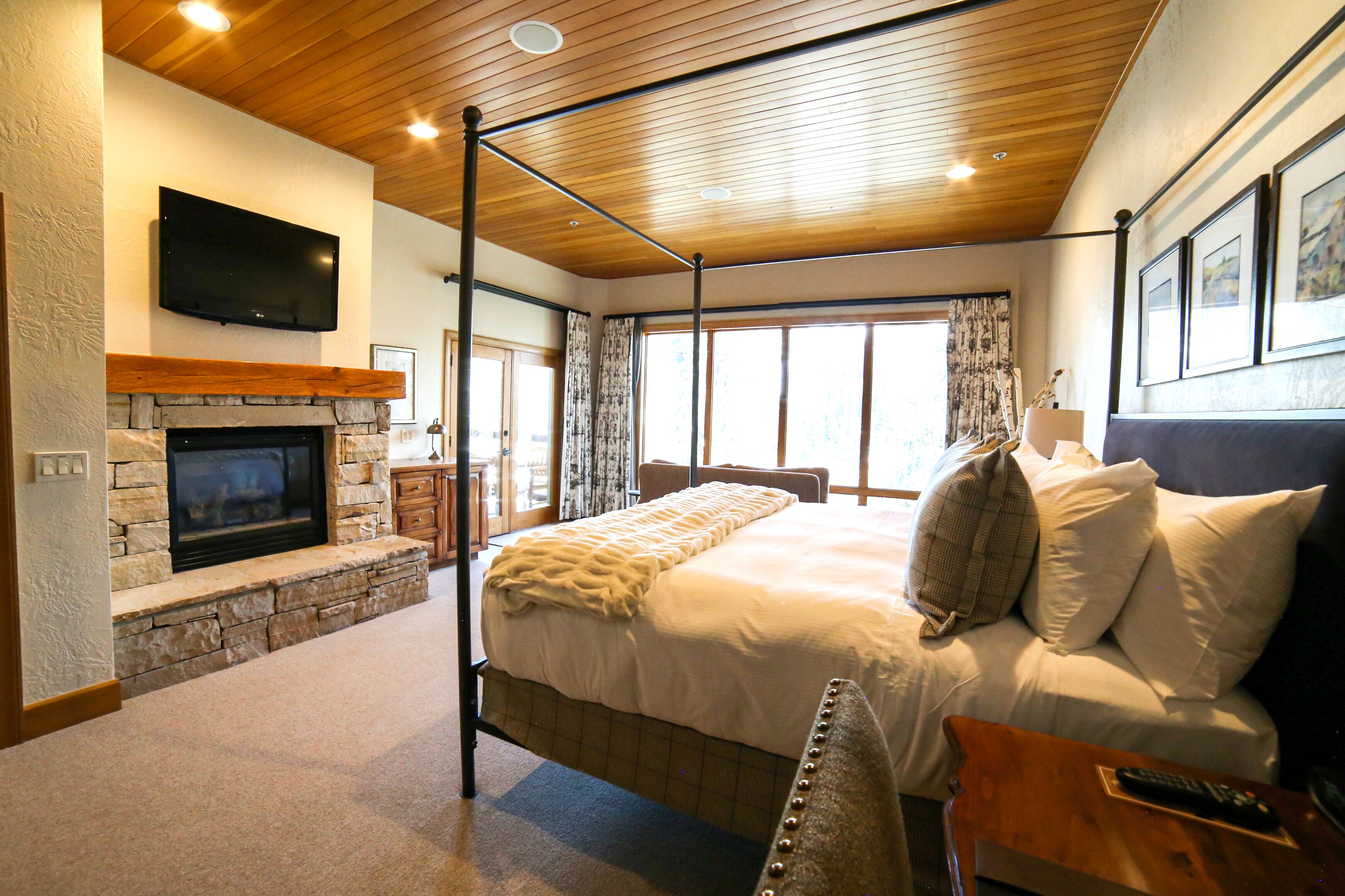 Un dormitorio con chimenea de piedra, una cama grande con ropa de cama blanca, techo de madera y televisor de pantalla plana montado en la pared.