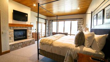 A bedroom with a stone fireplace, a large bed with white bedding, a wooden ceiling, and a flat-screen TV mounted on the wall.