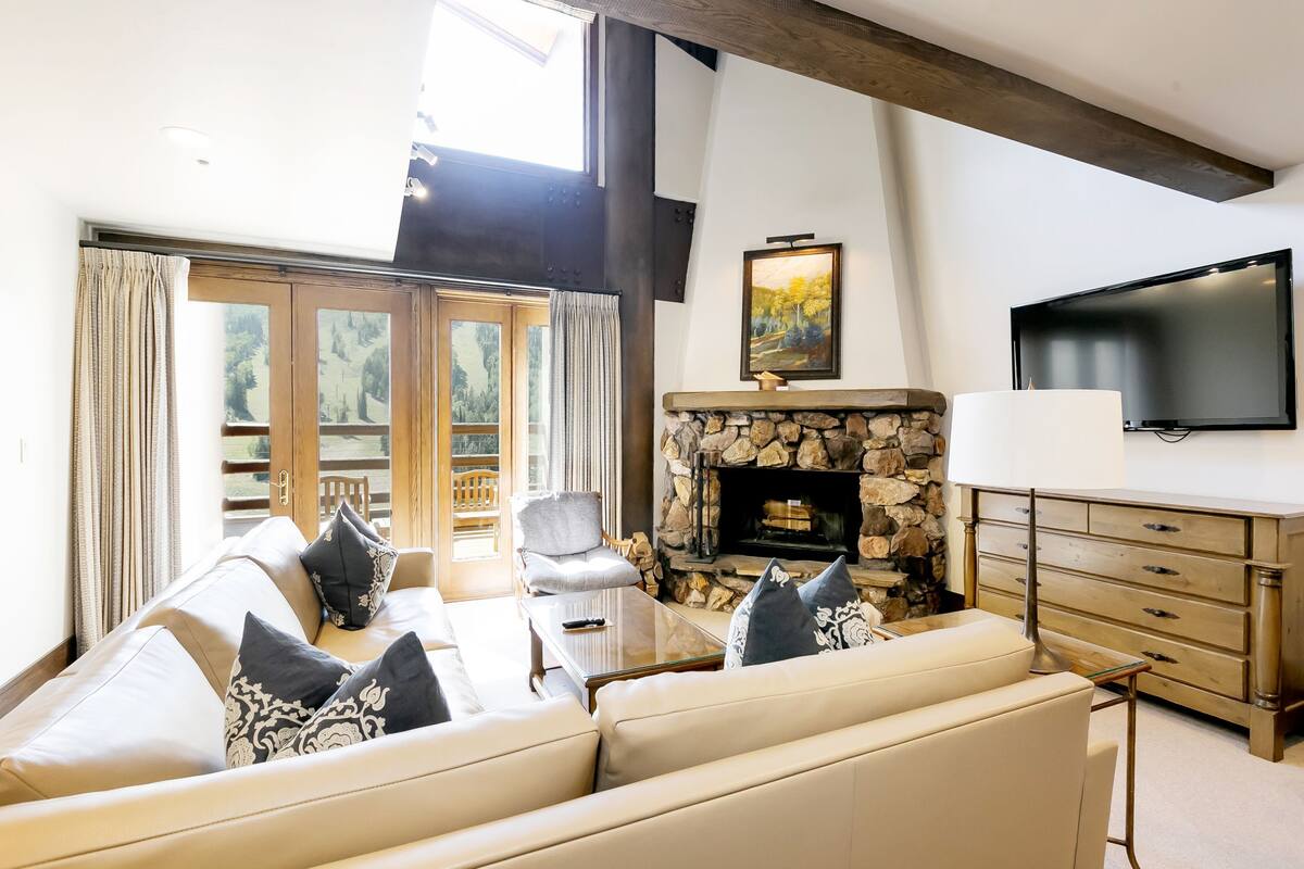 Ein Wohnzimmer mit Kamin aus Stein, einer Sofagarnitur, einem Fernseher und einem großen Fenster mit Vorhängen.