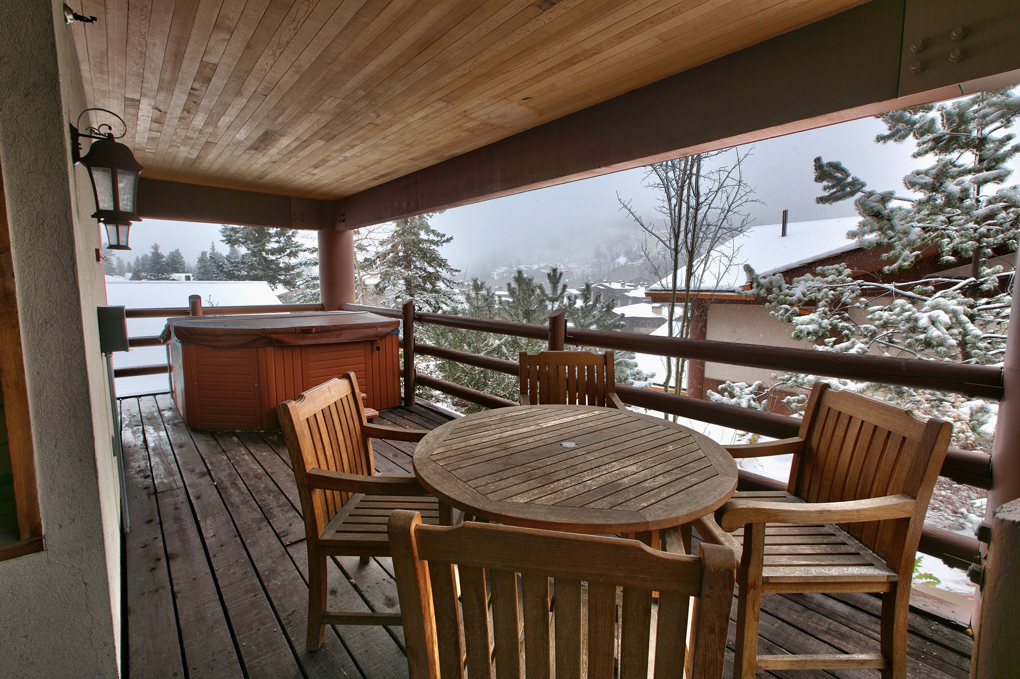 Une terrasse en bois avec une table ronde et des chaises, un jacuzzi et une vue sur des arbres et des bâtiments recouverts de neige.