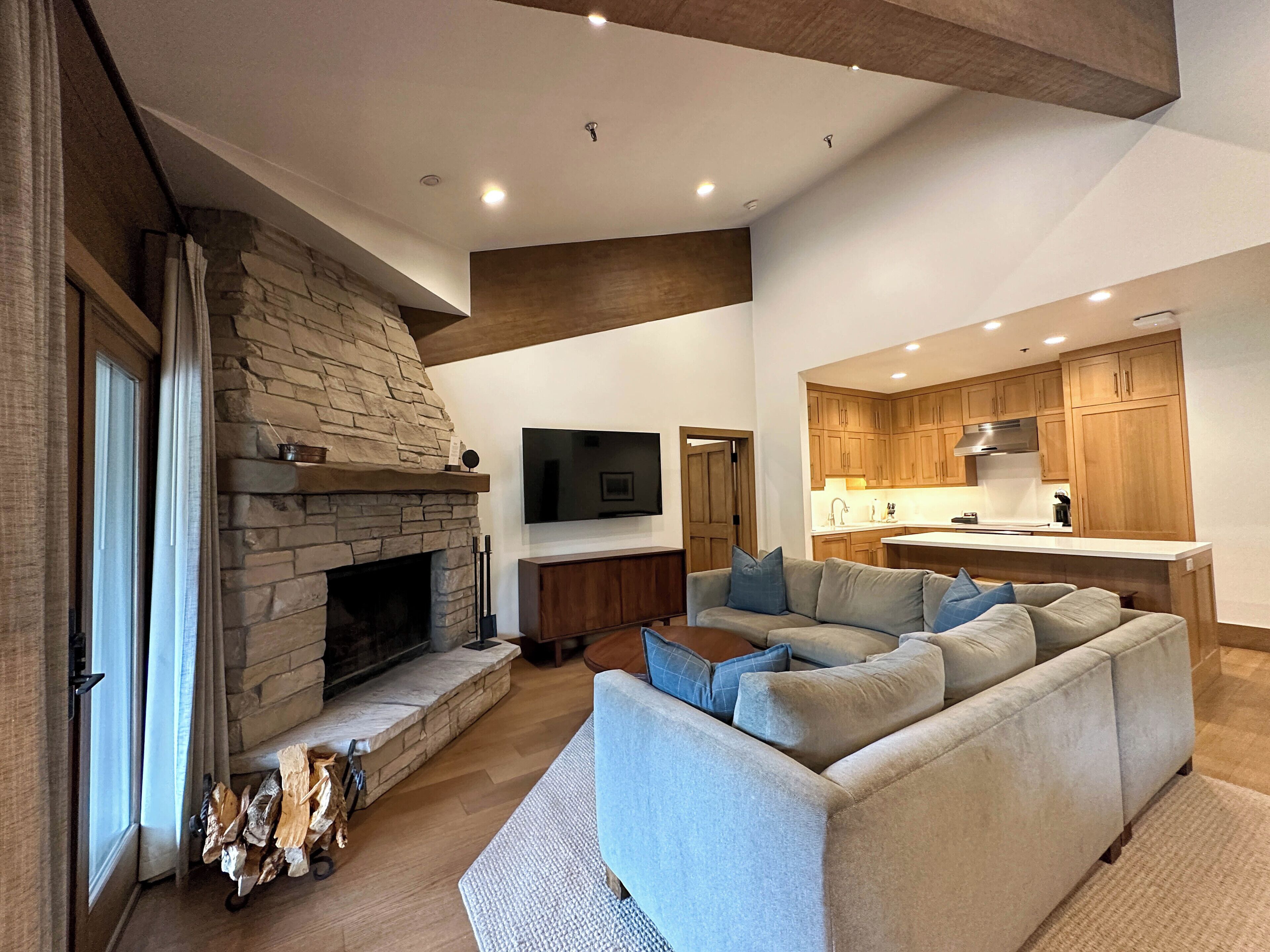 Ein modernes Wohnzimmer mit Kamin aus Stein, einem großen grauen Ecksofa und einer Küchenzeile mit Holzschränken sowie einem Flachbildfernseher.