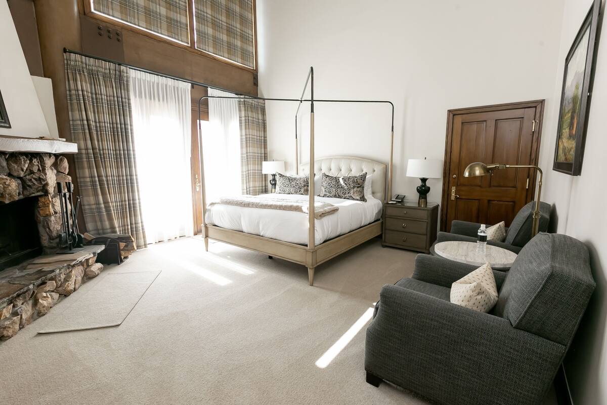 Ein Schlafzimmer mit Himmelbett, grauem Sessel, rundem Tisch und Holztür.