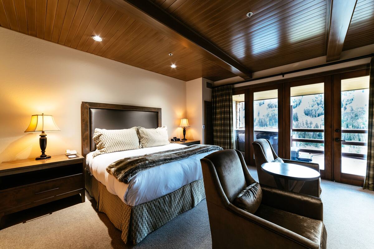 Ein Hotelzimmer mit einem großen Bett, zwei Nachttischlampen, einem Sessel, einem kleinen Tisch und einem Balkon mit Blick auf Bäume.