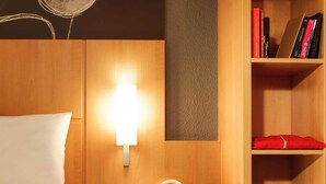 A hotel room with a bed, bedside table with a tablet and headphones, a wall-mounted lamp, and a bookshelf with books and a folded red garment. - ibis Epinay sur seine Gennevilliers (Epinay-sur-Seine)