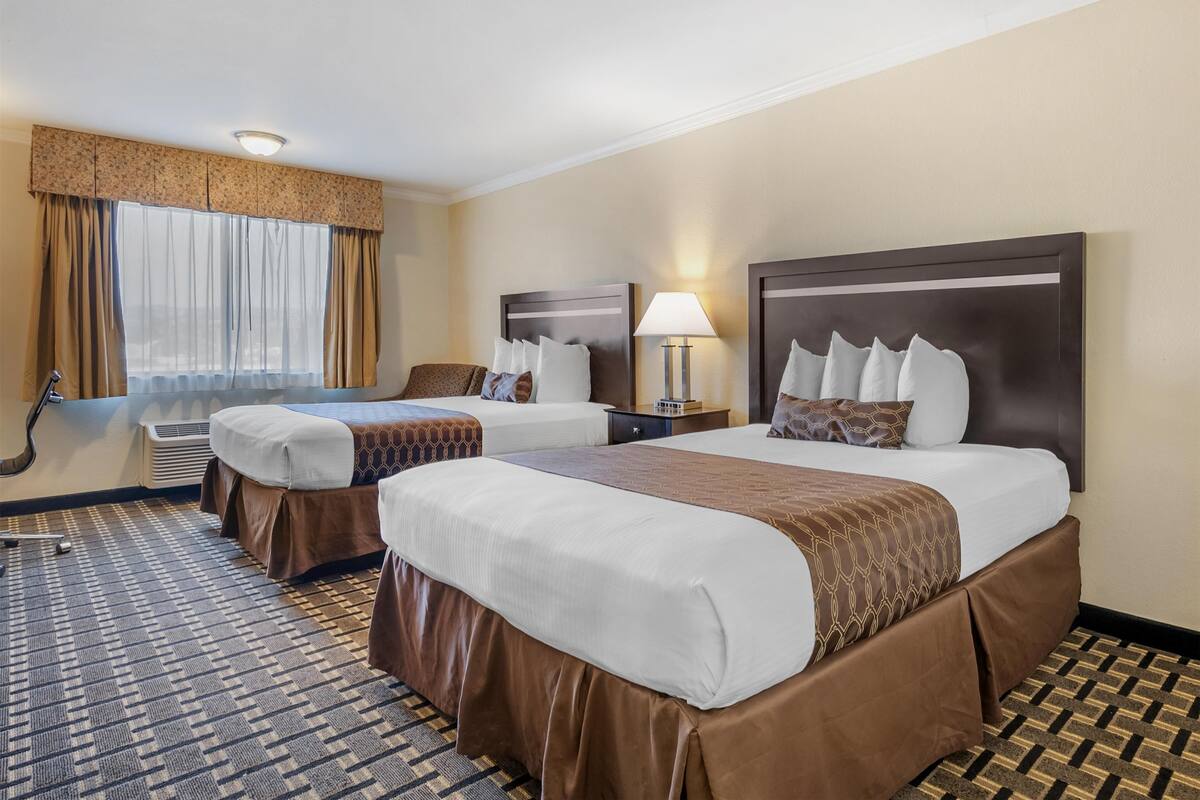 Une chambre d’hôtel avec deux lits, une grande fenêtre avec des rideaux, une table de chevet avec une lampe et une moquette à motifs.
