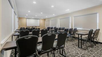A conference room with rows of chairs and tables, a projector screen, and large windows with blinds.