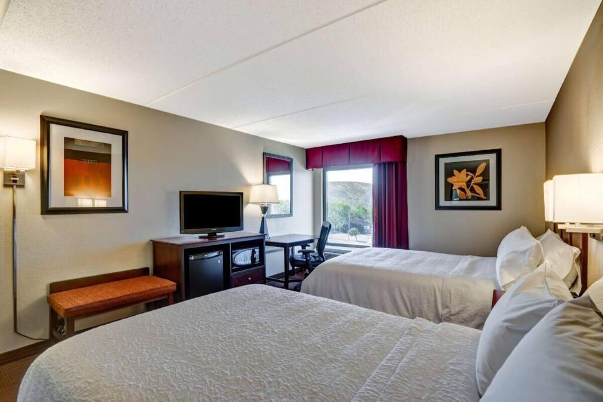 A hotel room with two beds, a desk with a television, a bench, and framed artwork on the walls.