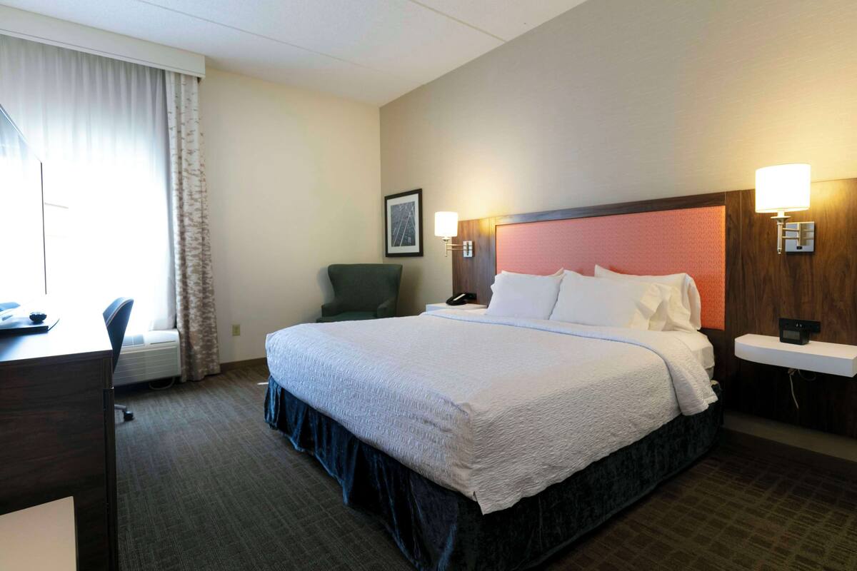 A hotel room with a large bed, a desk, a chair, and a bedside table with a lamp.