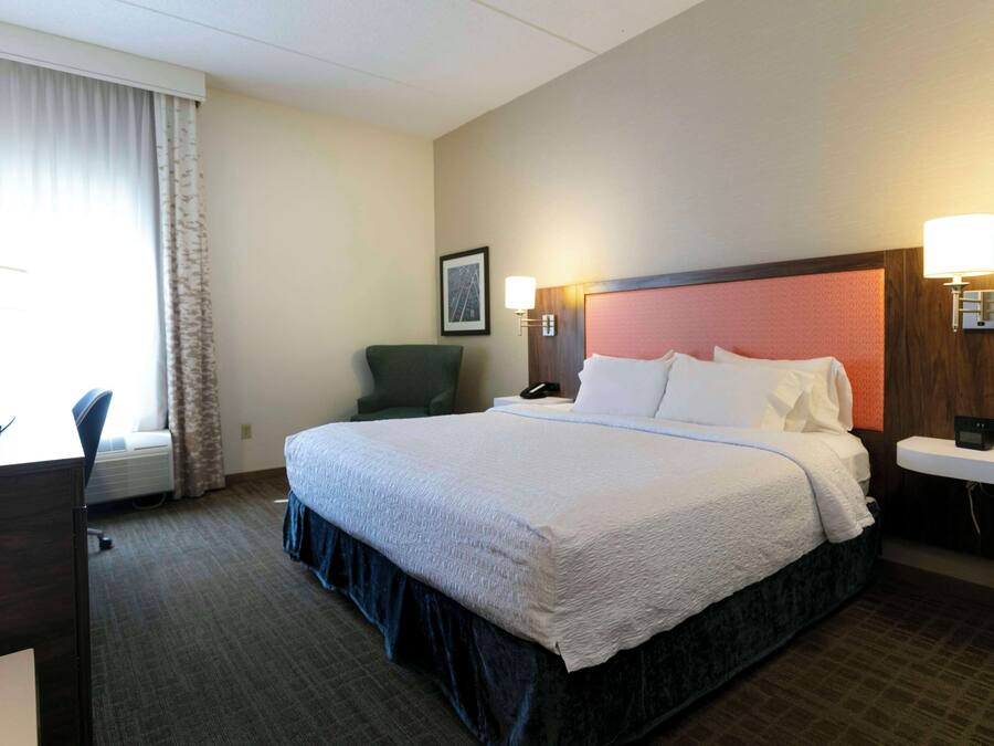A hotel room with a large bed, a desk, a chair, and a bedside table with a lamp.