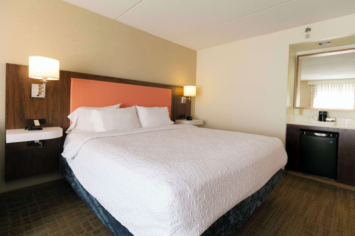 A hotel room with a large bed, two bedside tables, a built-in desk with a lamp, a mirror, and a small refrigerator.
