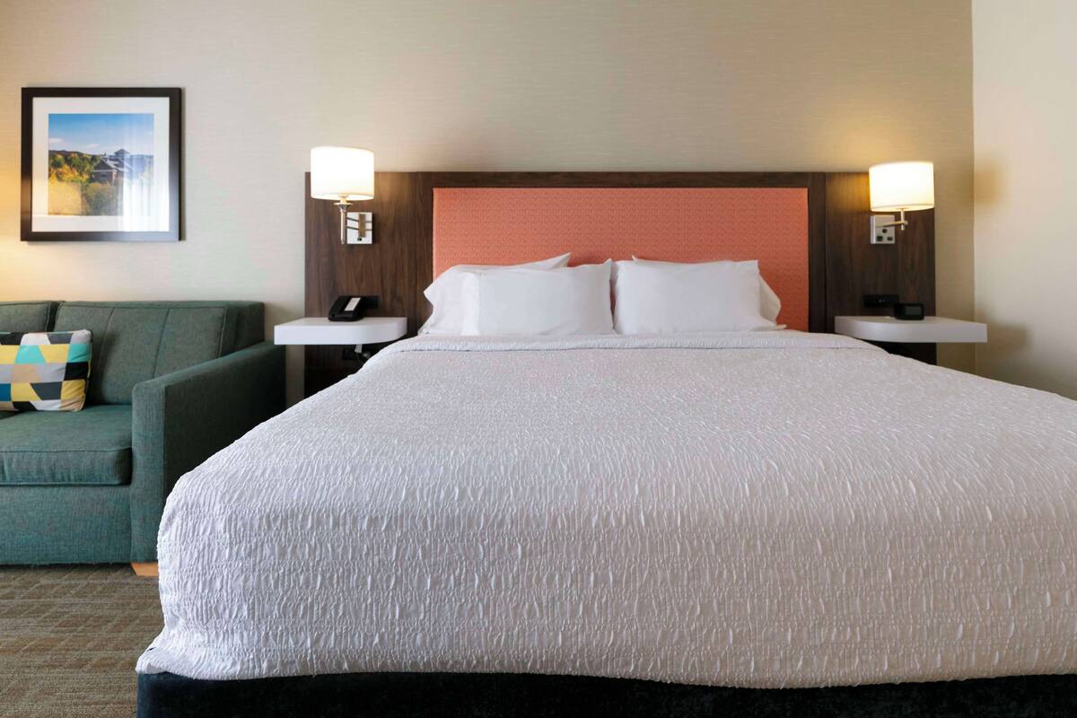 A hotel room with a large bed, two bedside tables with lamps, a green armchair, and a framed picture on the wall.