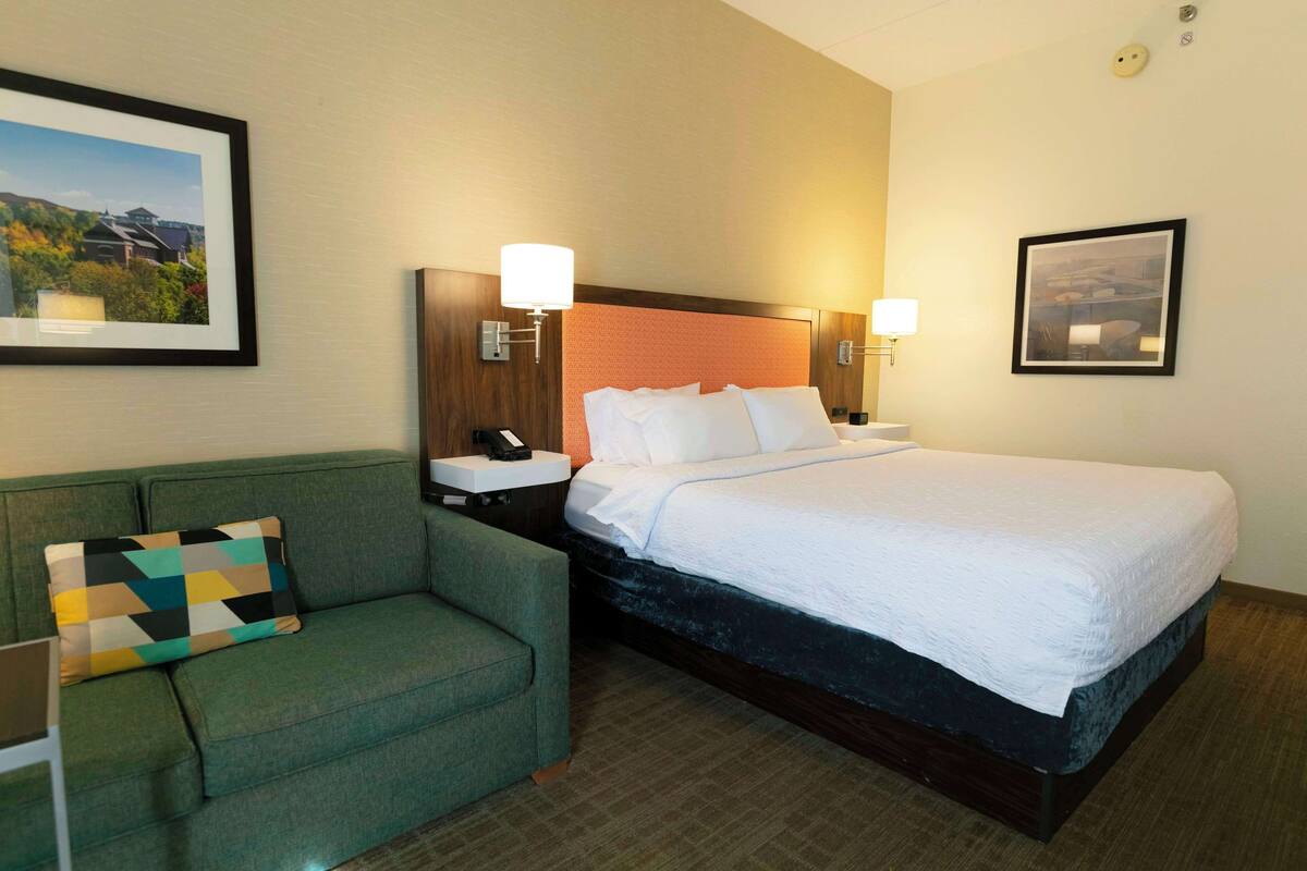 A hotel room with a bed, a green sofa, a nightstand with a lamp, and framed pictures on the walls.