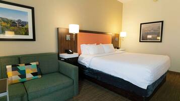 Habitación de hotel con cama, sofá verde, mesita de noche con lámpara y cuadros en las paredes.
