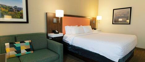 Habitación de hotel con cama, sofá verde, mesita de noche con lámpara y cuadros en las paredes.