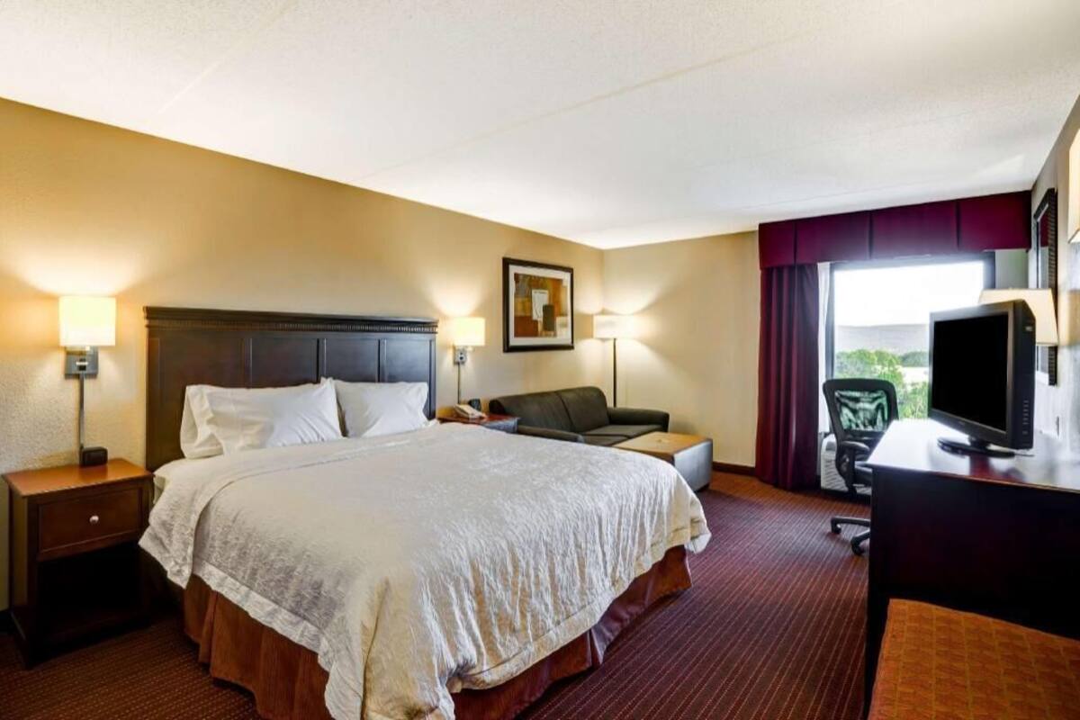 A hotel room with a large bed, a desk with a computer, a chair, and a window with curtains.