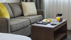 A modern living room with a grey sofa, a wooden coffee table, and a breakfast setup including eggs benedict, orange juice, and a coffee pot. - Holiday Inn Winnipeg South by IHG (Winnipeg)