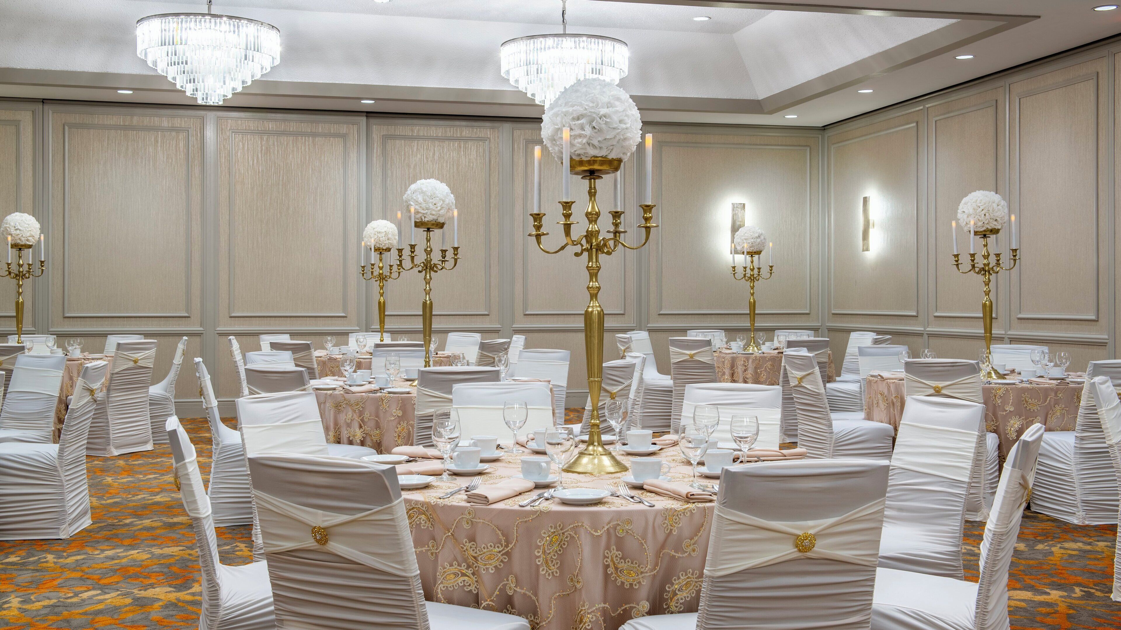 Salão de banquetes com mesas redondas, cadeiras brancas e candelabros dourados.