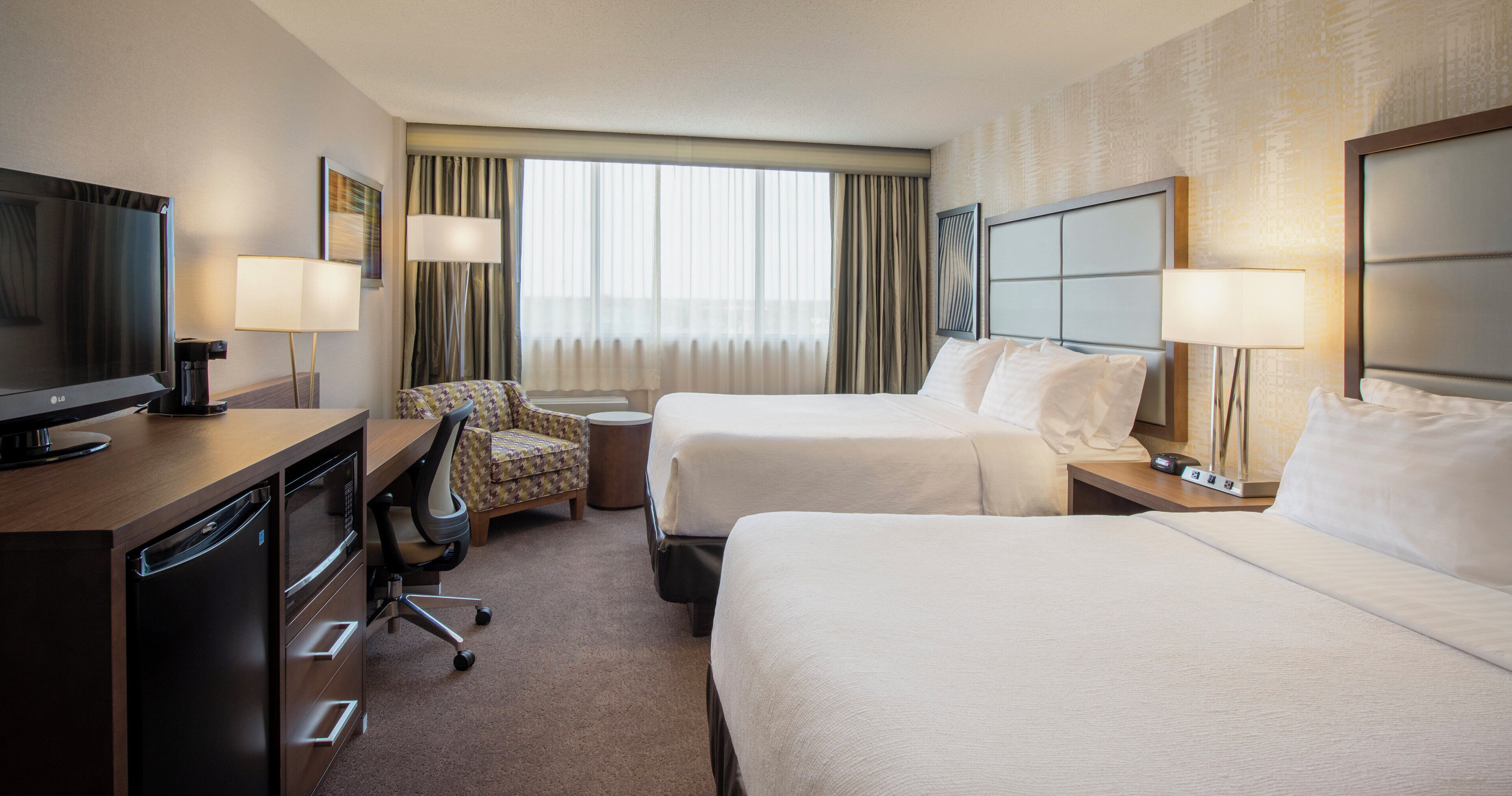 A hotel room with a bed, a desk with a television, a chair, and a window with curtains.